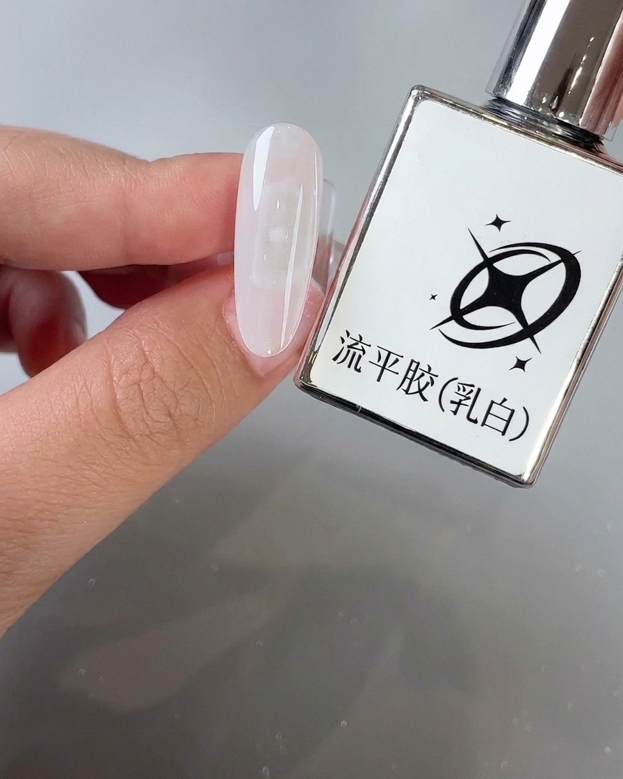 製品ボトルの隣で、アーモンド型のネイル表面にブラシでミルキーホワイトのレベリングジェルを塗っている様子。