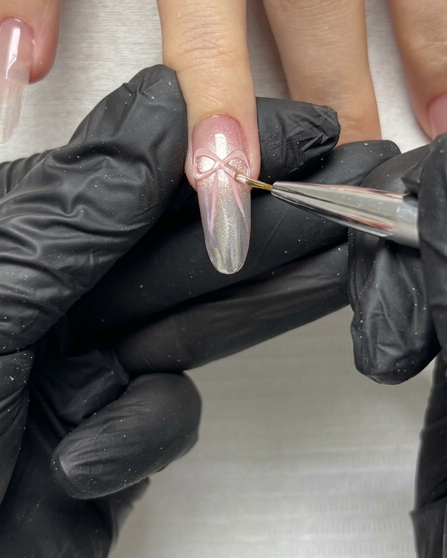 マニキュアを施した爪の上のピンクの3Dリボンボウの繊細なエッジやテールを、細いブラシで微調整しているクローズアップショット。