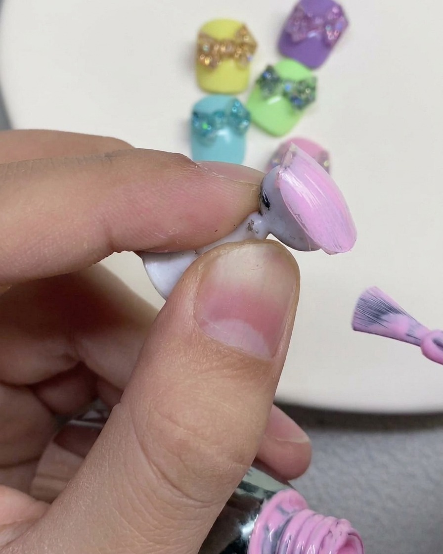 ネイルホルダーに固定されたフェイクネイルの表面に、ネイルブラシでソフトなパステルピンクのジェルポリッシュを塗布している様子。