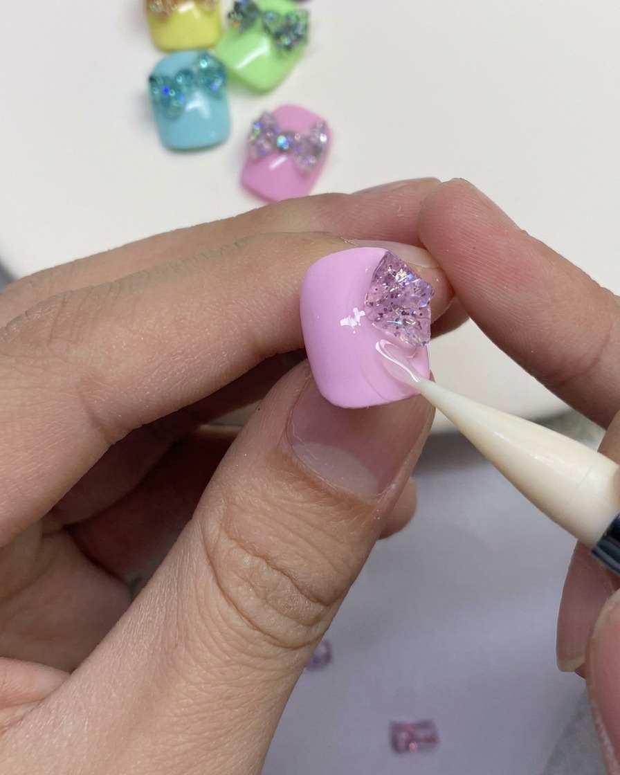 ピンクのネイルの上で、シリコンツールを使ってグリッタージェルのパーツをバタフライリボンの外側ループへと形作っているクローズアップ。