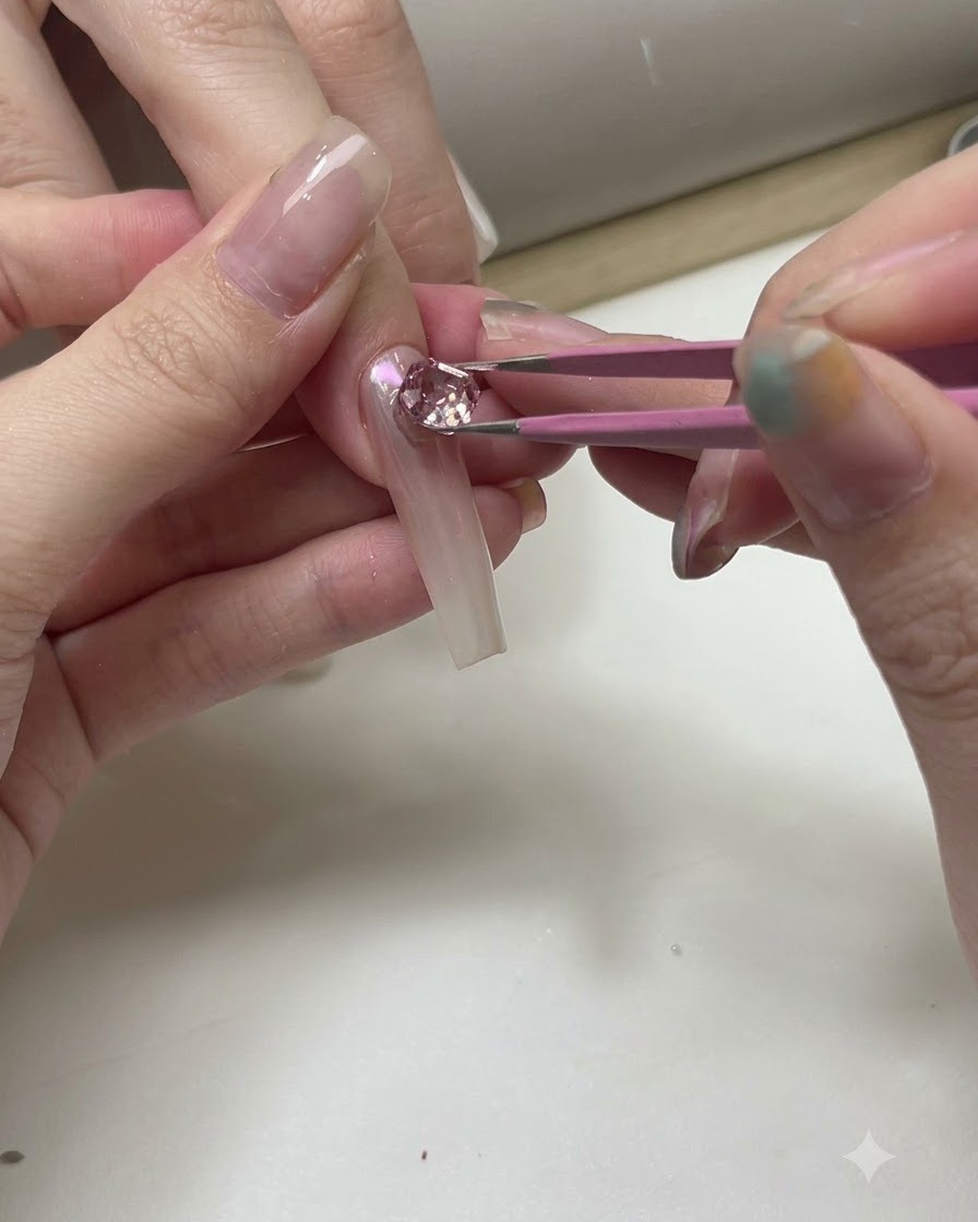 ピンセットを使って、透明な練習用ネイルチップの上にスパークリングピンクやクリアのクリスタルを配置している様子。