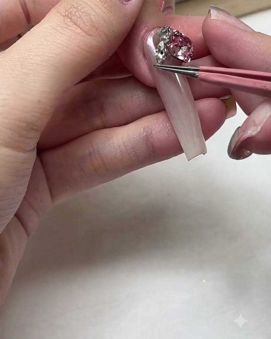 ピンクのピンセットがロングなネイルベッドの上に輝く大粒のピンククリスタルラインストーンを置いている様子。