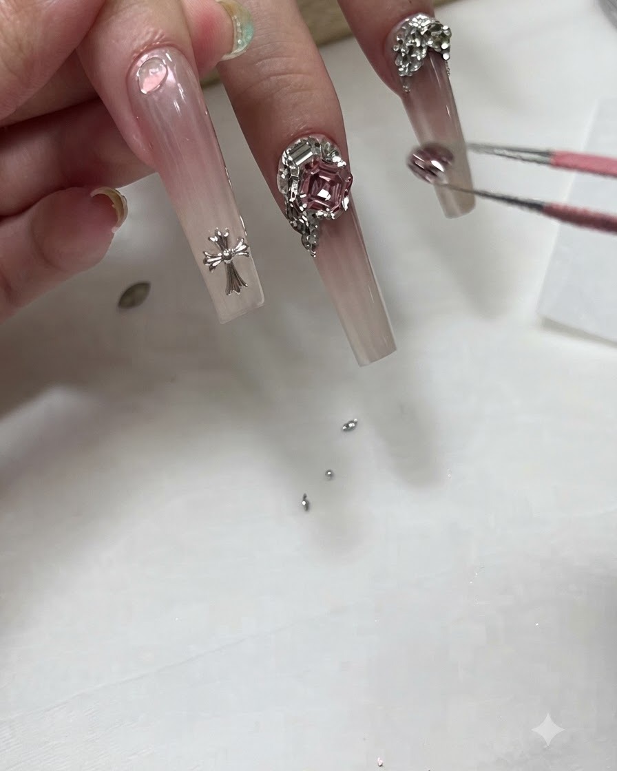 ネイリストがピンセットを使って、ロングなオンブレネイルの根元の近くに小さなクリスタルチャームを配置している様子。