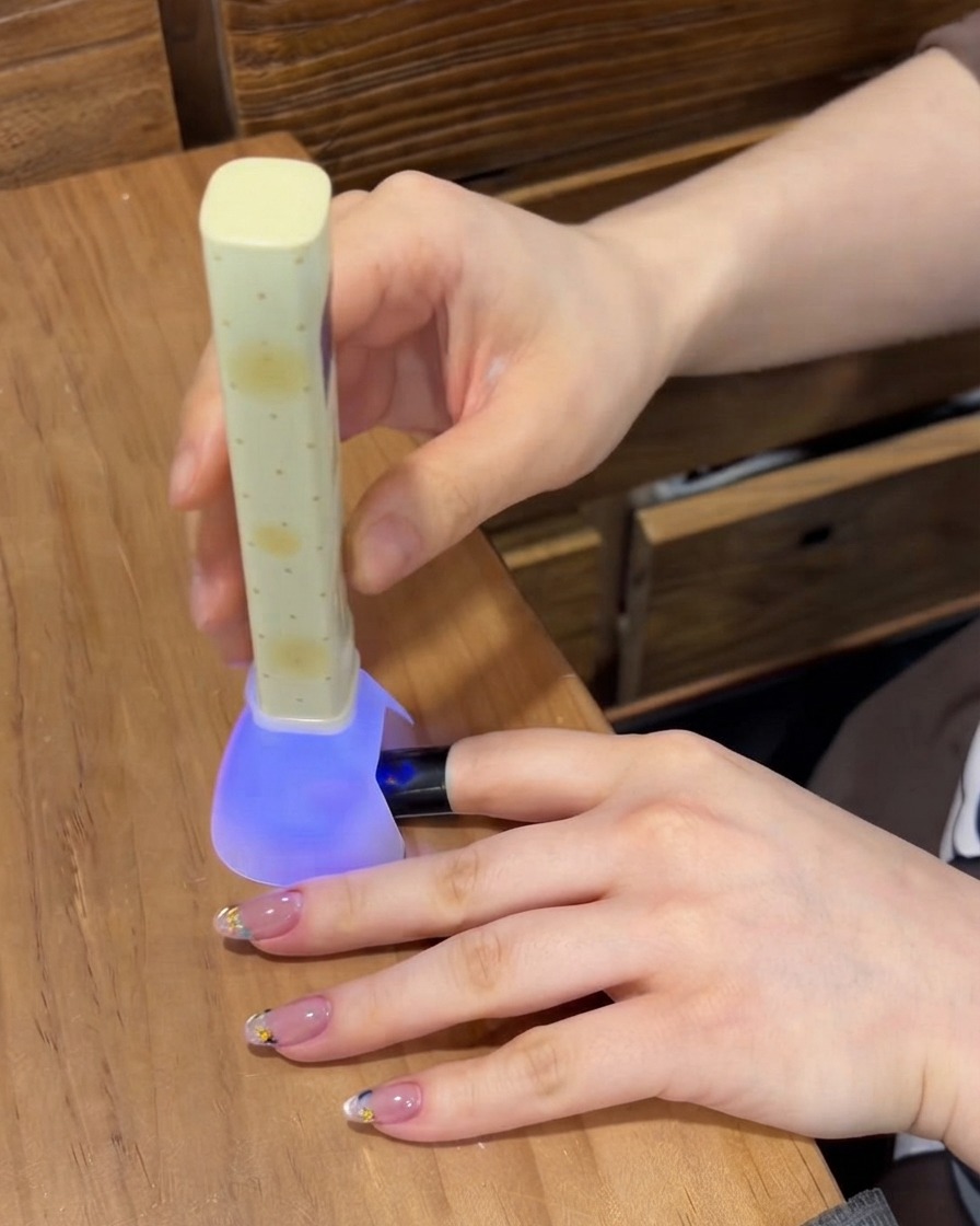 ポータブルな紫色のUV/LEDネイルランプの下で爪の上のジェルを硬化させている様子。テーブルには他の整えられた指が置かれている。