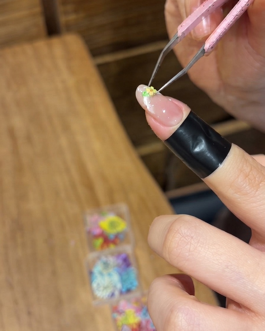 精密ピンセットを使って、指の爪の濡れたジェル表面に小さな黄色いドライフラワーを配置している様子。