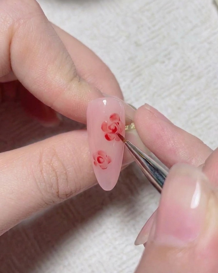 ピンクのネイル上に描かれたバラの中心を形作るために赤いディテールラインを追加している細いネイルアートブラシ。