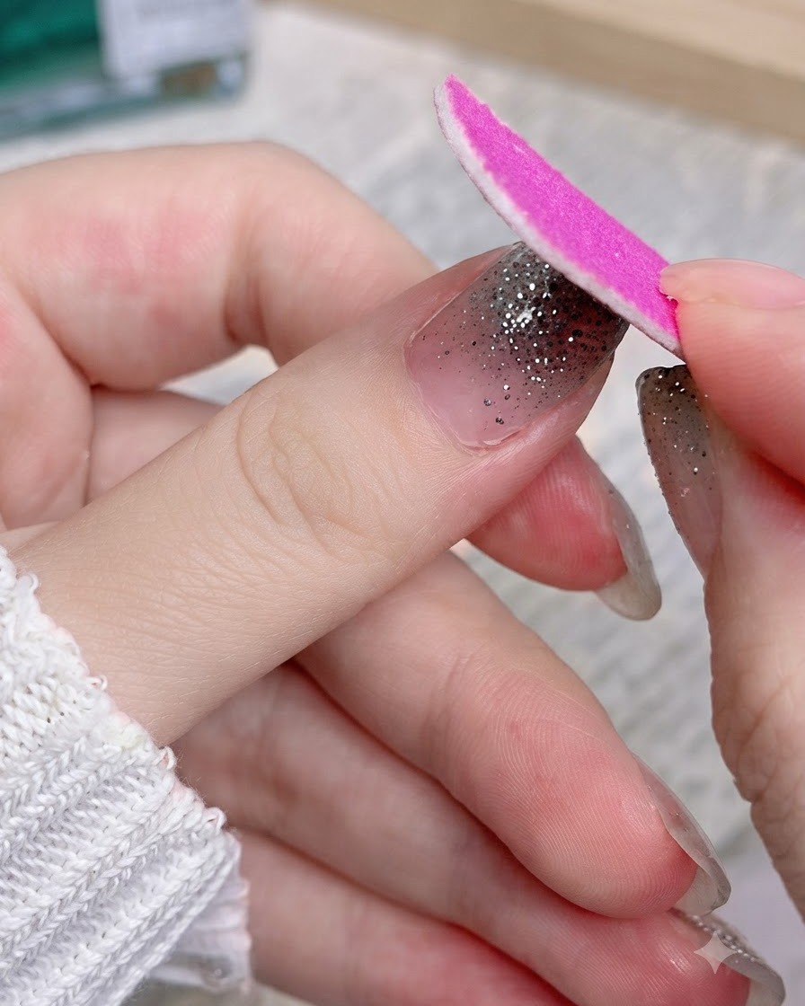 ピンクのエメリーボードを使い、親指のシルバーラメネイルシールの余分な部分をファイルで削り落としている様子。