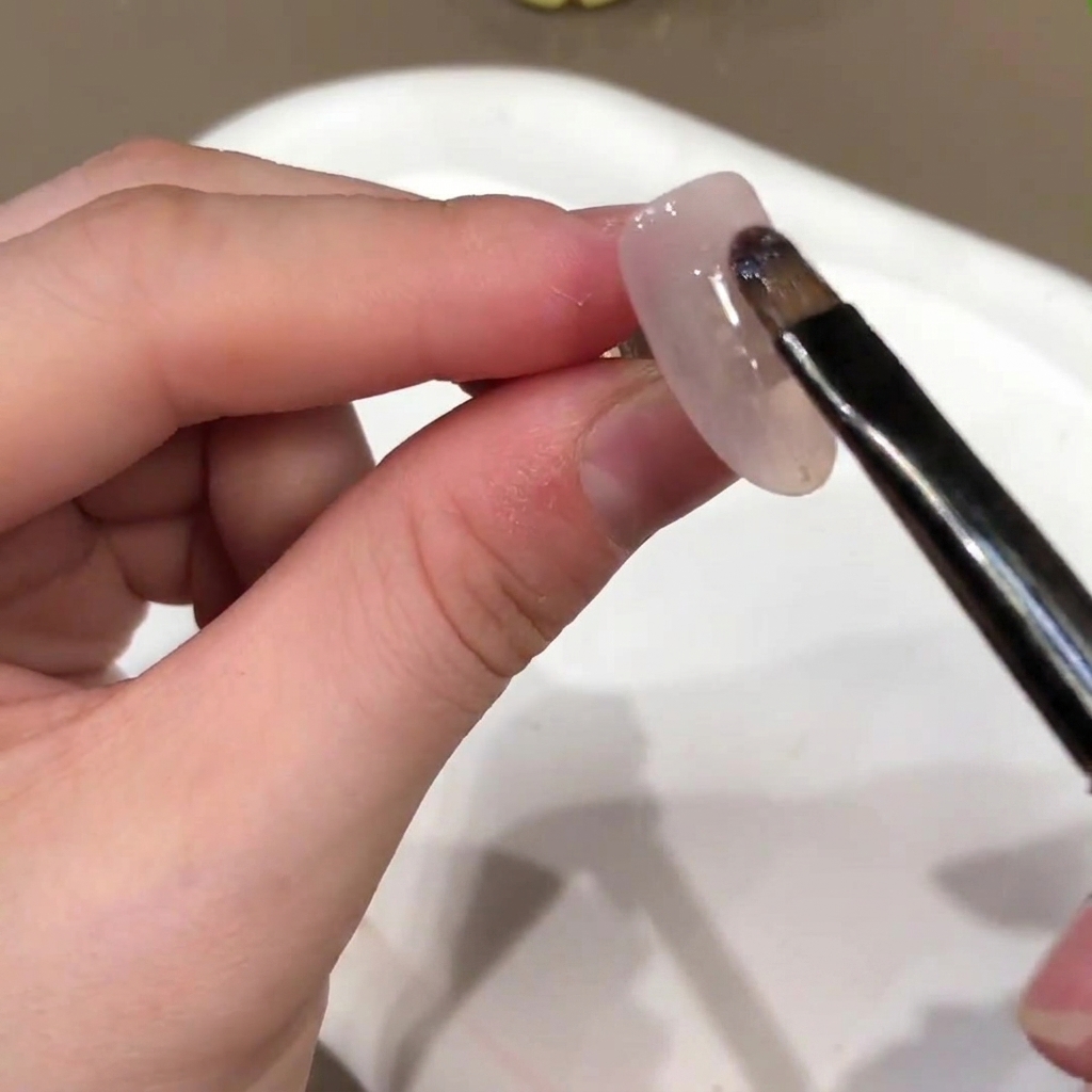 ネイルブラシが人工爪の表面にヌードピンクのジェルベースカラーを塗布している様子。