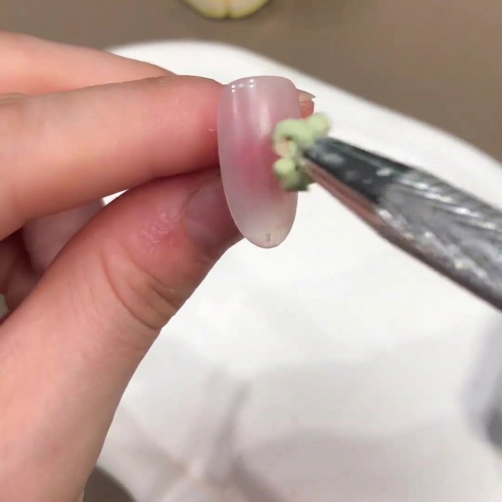 小さなブラシが、ヌードピンクのネイルの中央にグリーン系ジェルの小さな塊を載せている様子。