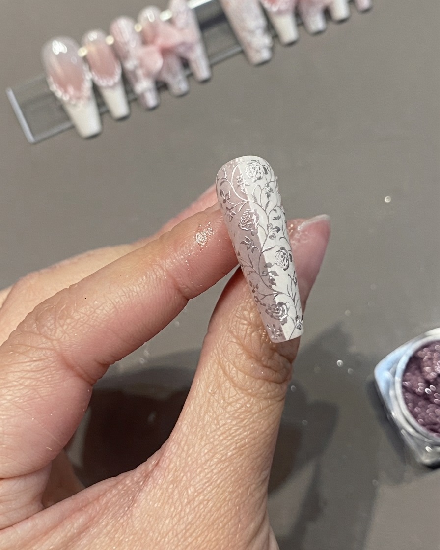 指の間に持たれた、詳細な白いフローラルレースとクロムパターンを特徴とするロングコフィンネイルチップのクローズアップ。
