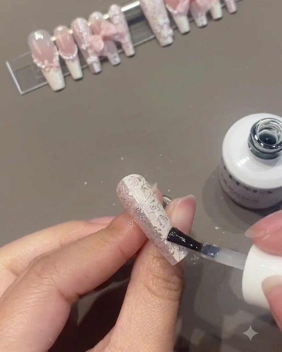 背景に他の完成したネイルがディスプレイスタンドに置かれている中、複雑に装飾されたフローラルネイルの上にクリアまたはトップコートを塗布しているネイルブラシ。