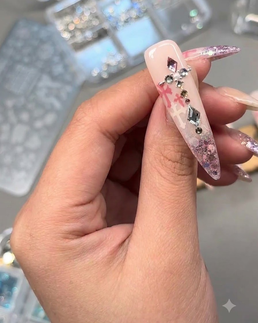 ピンクのグリッターとスタンプアートの上に、クリスタル、パール、メタルチャームが飾られたネイルのクローズアップ。