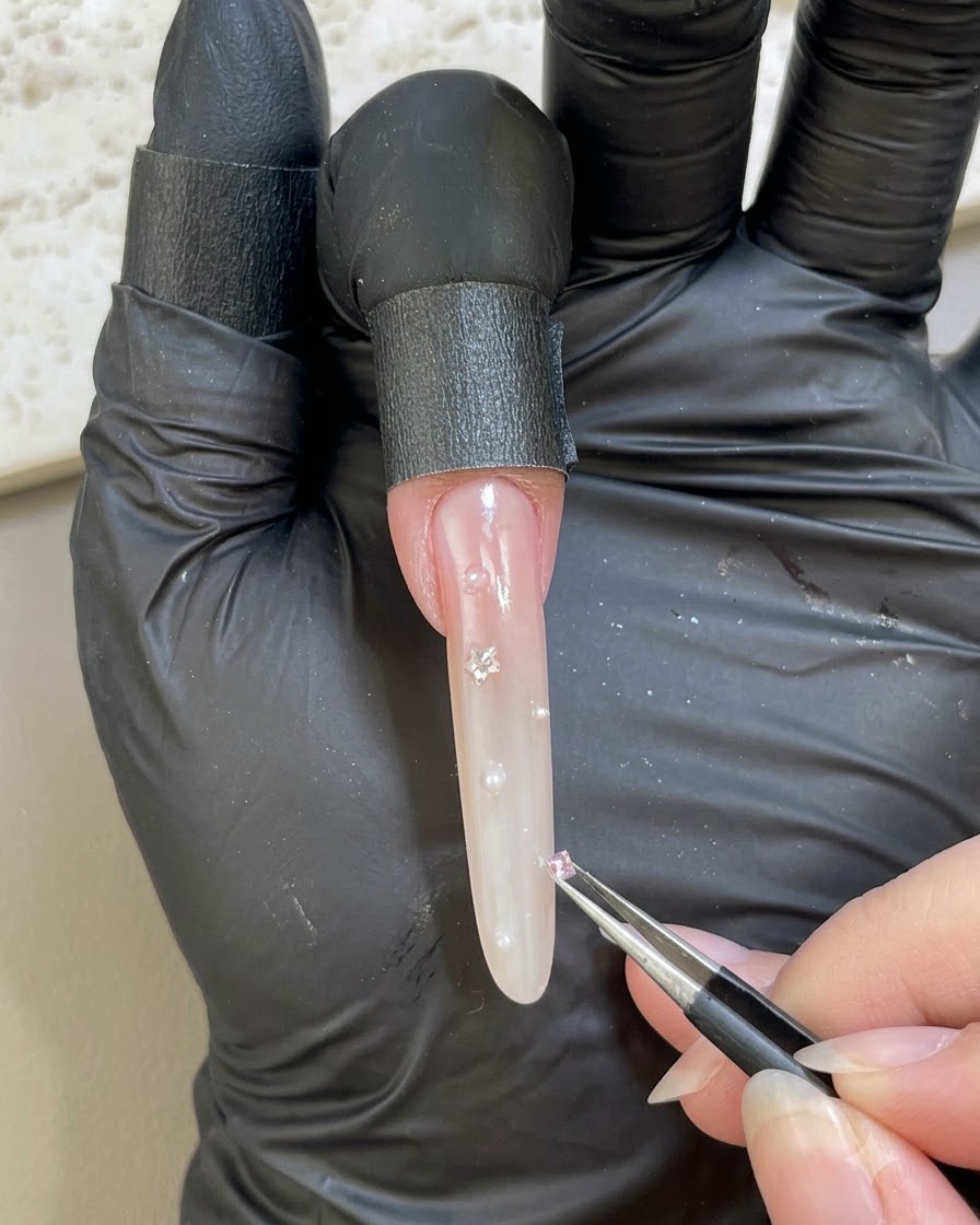 手袋をした手がピンセットを使って装飾されたネイル表面に小さなピンクのラインストーンを置いている様子。