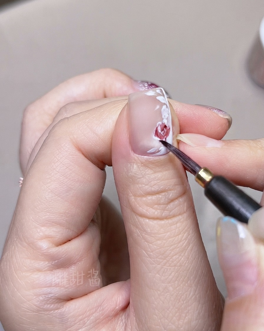 細いブラシを使って、爪の上に描かれたバラの花びらや葉に濃い赤の水彩トーンを重ねている様子。