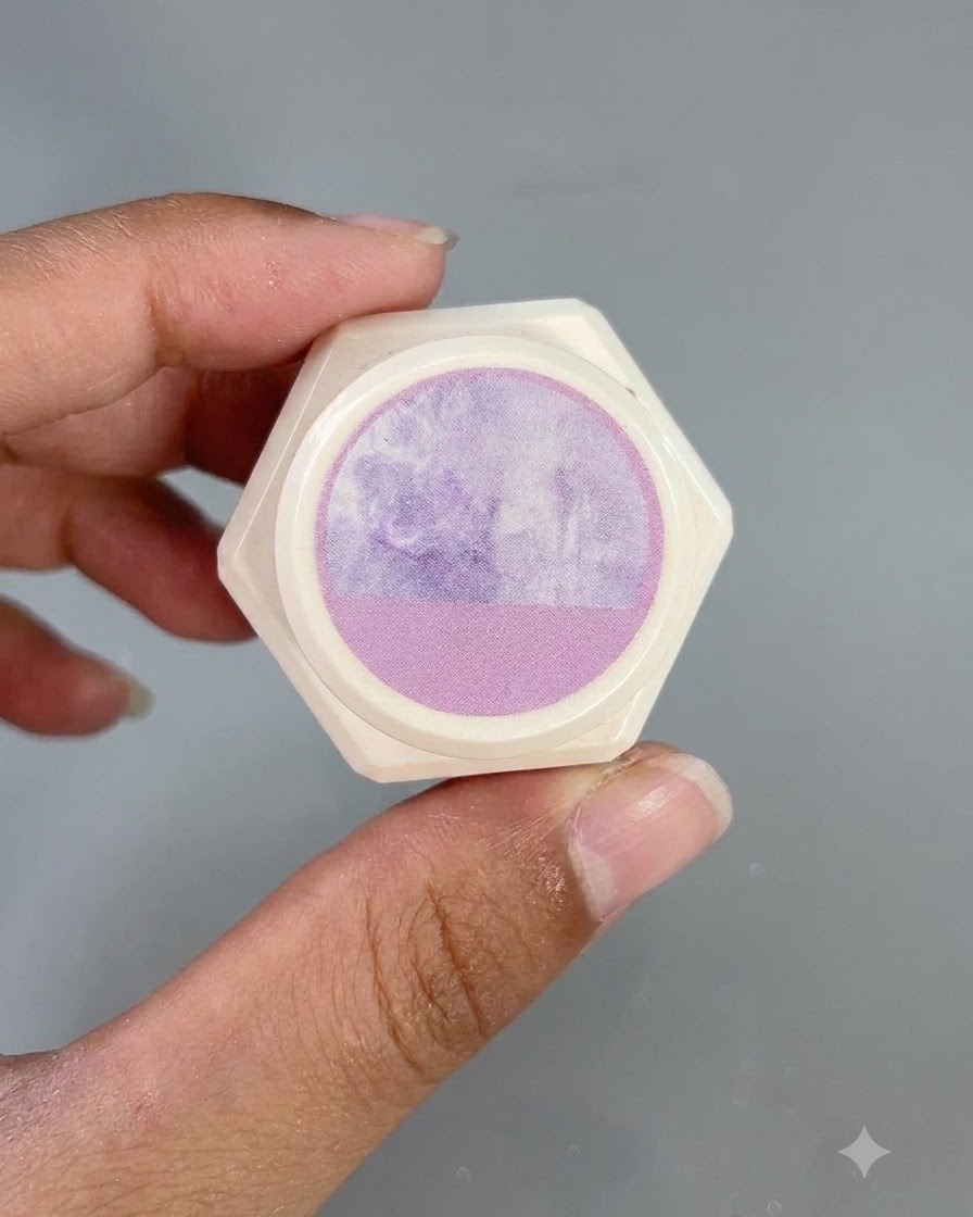 指で持ち上げられた、蓋にパープルの水彩パターンのステッカーが貼られた六角形の白い容器。