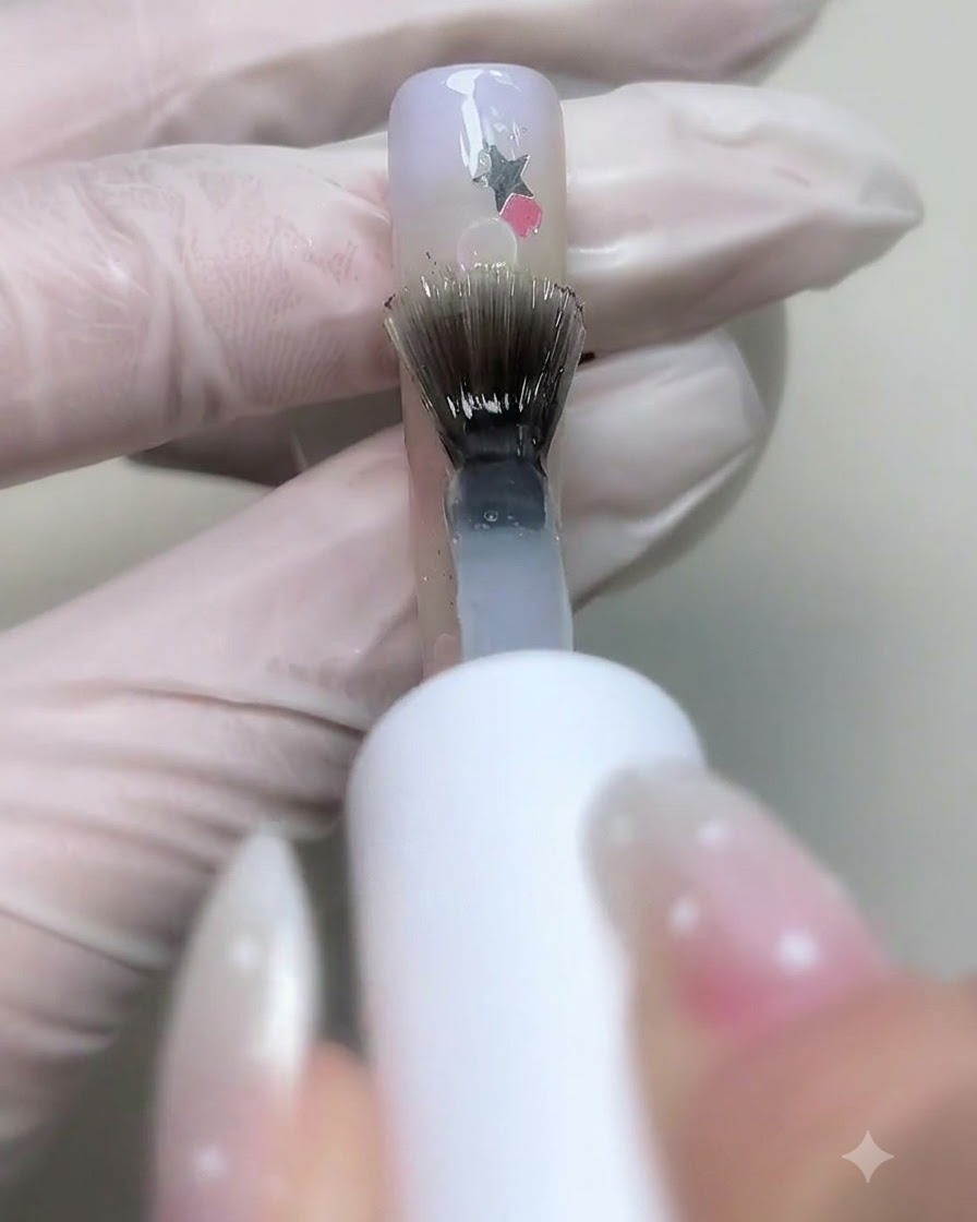 スターグリッターとバブルで装飾されたネイルの上に、クリアベースジェルをブラシで塗布している様子。
