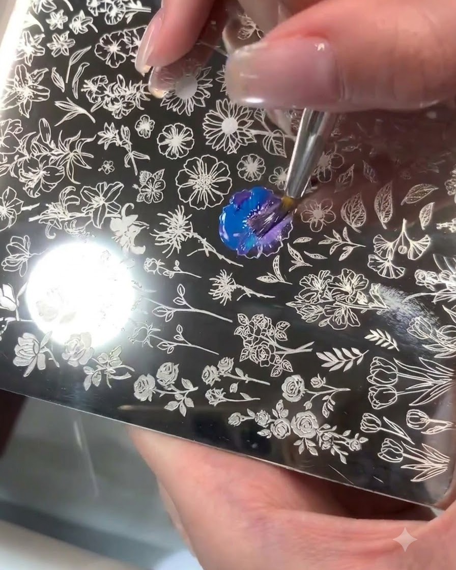 彫刻された花の模様が施されたスティール製スタンピングプレート上で、ブラシで青と紫のジェルを混ぜ合わせている様子。