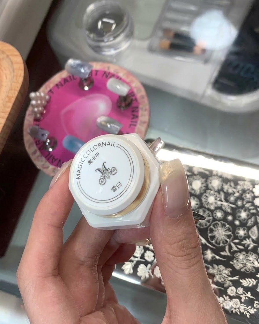 背景にネイルスタンピングプレートを置き、アジア文字のラベルが貼られた白いボトルのスタンピング製品を持っている手。