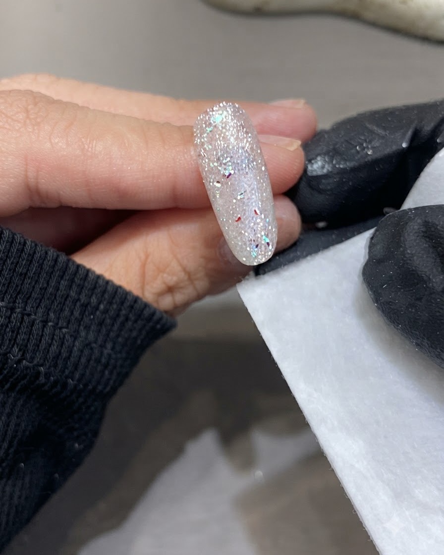 硬化させたきらめくネイルの隣で、手袋をした手がリントフリーワイプを持っている様子。