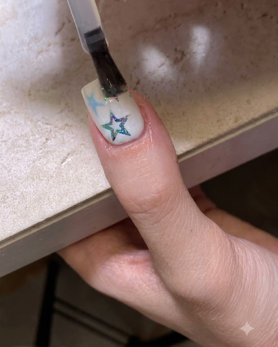 星とグリッターで装飾された爪の上に、ブラシで光沢のあるクリアトップコートを塗布している様子。