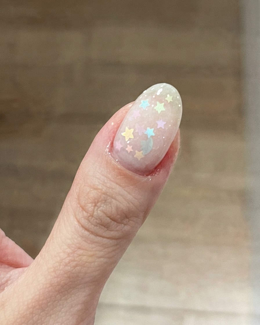ミルキーピンクのベースの上に、カラフルなパステルカラーの星型グリッターが配置された親指のクローズアップ。