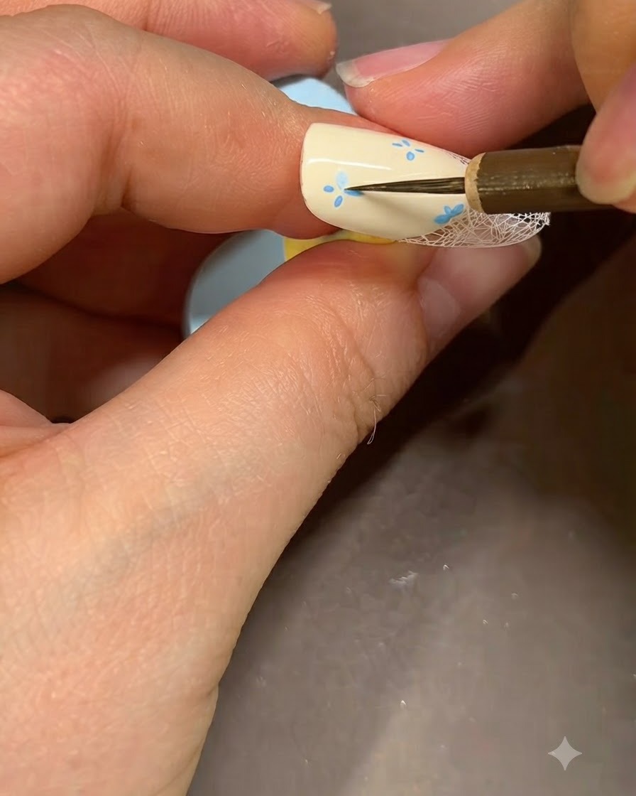 極細のネイルアートブラシを使い、ベージュのネイルに描いた花の中心に細かいブルーのアクセントを加えている様子。