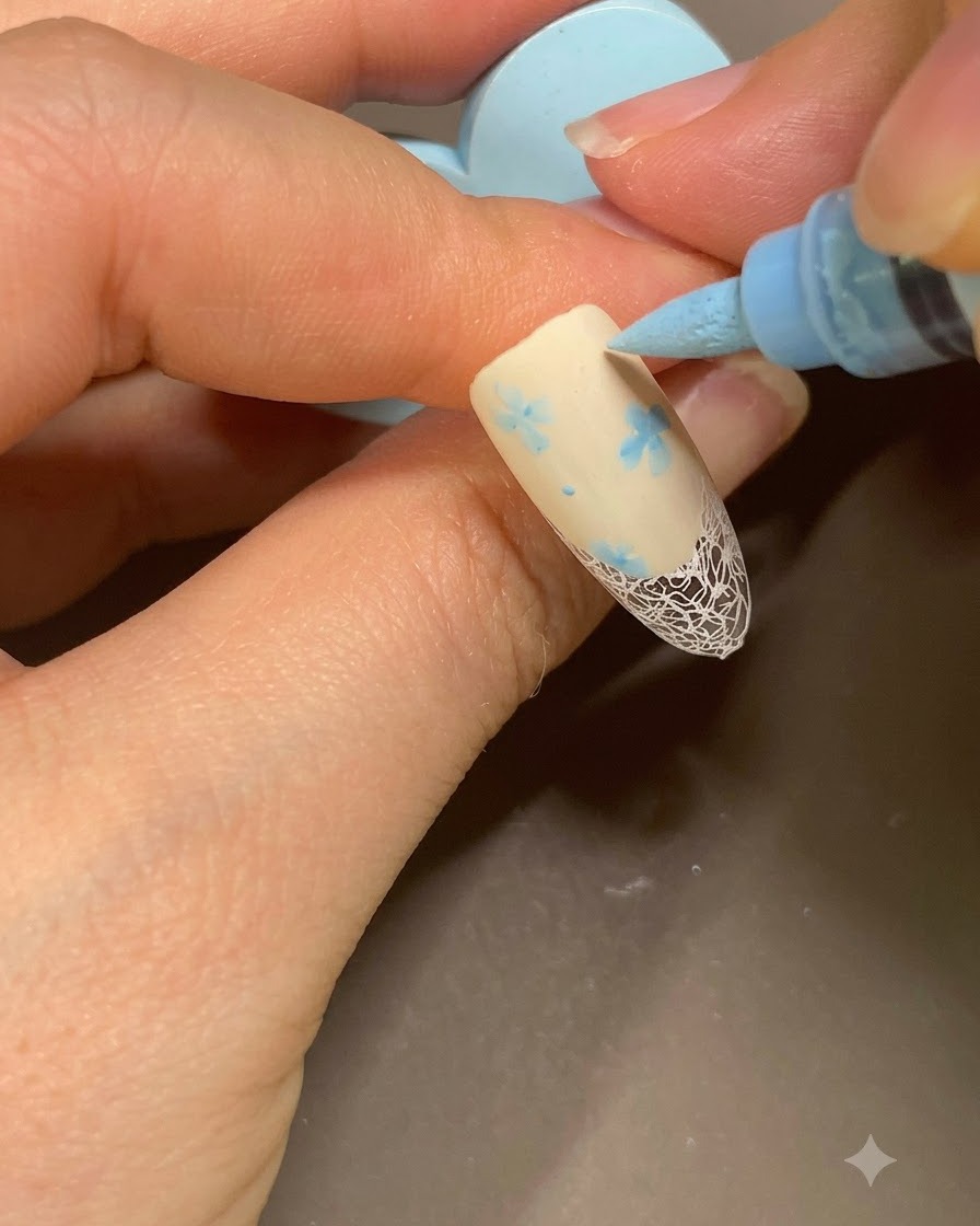 ベージュのフレンチマニキュアの花柄の横に、青いネイルアートマーカーで小さなドットを追加している様子。
