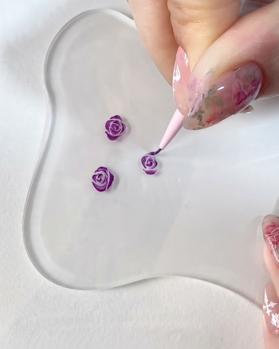 クリアなガラスパレット上で、手持ちの細いディテールブラシを使って小さな3Dローズのデザインに濃い紫色のアクセントを細かく描き込んでいるクローズアップ画像。