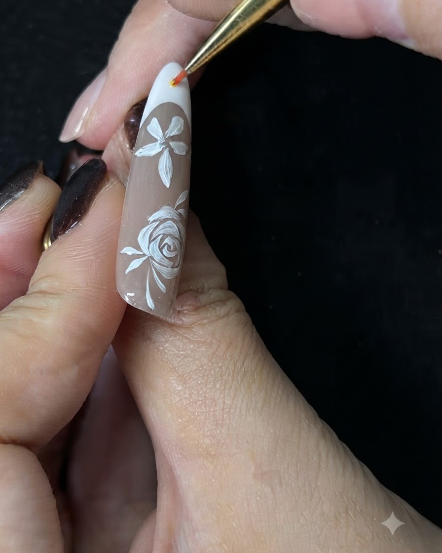 ヌードジェルのネイル上に手描きされた白い薔薇の周りに、極細のディテールブラシで小さな白いアクセントのツタを描いている手のクローズアップ。
