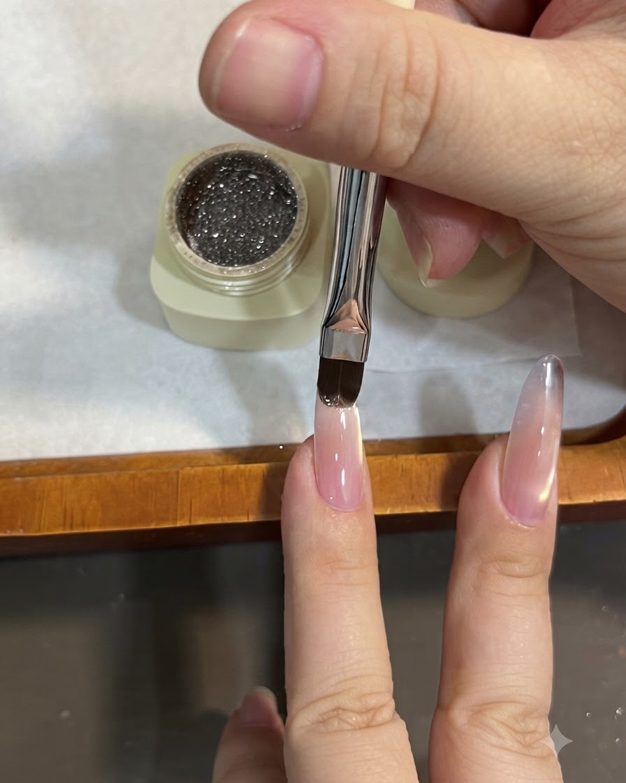 フラットブラシを使ってアーモンドネイルの先端にスパークリングブラウンのグリッタージェルを塗り、オンブレ効果を生み出すために下方へ撫でつけている様子。