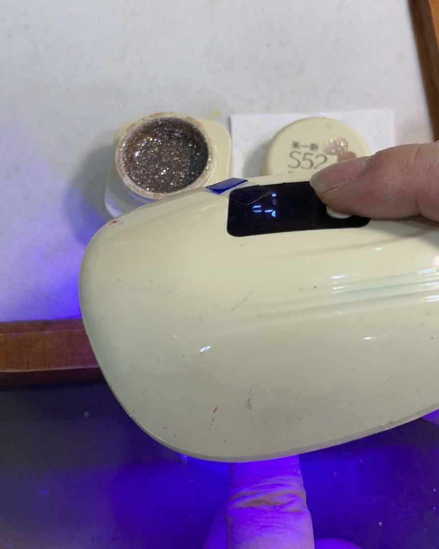 グリッターグラデーションが施されたネイルの指が、小型のポータブルUV/LEDネイルランプの下で硬化している様子。
