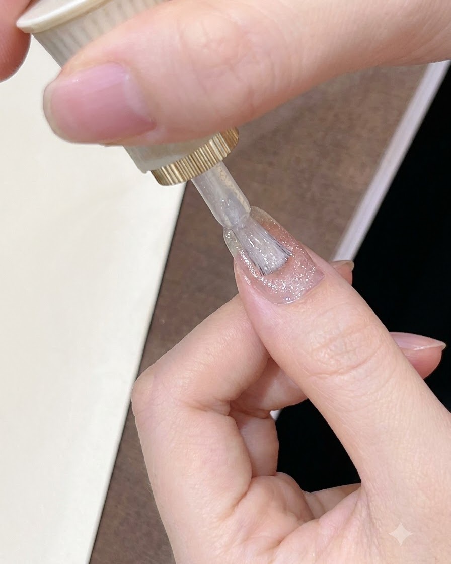 お手入れされた爪全体に、ブラシで透け感のある輝くピンクブラウンのジェルポリッシュを塗っている様子。
