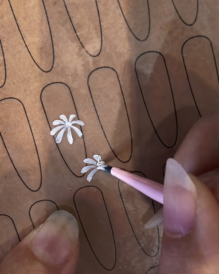 練習用のネイルシート上で、極細ライナーブラシが2つ目の小さめの白い花びらしを描き始めている様子。