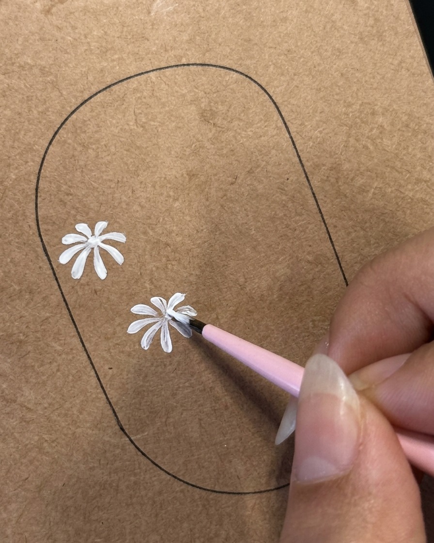 茶色のネイル練習用テンプレートに描かれた2つ目のデイジーのデザインで、極細ライナーブラシが白い花びらのディテールを描いている様子。