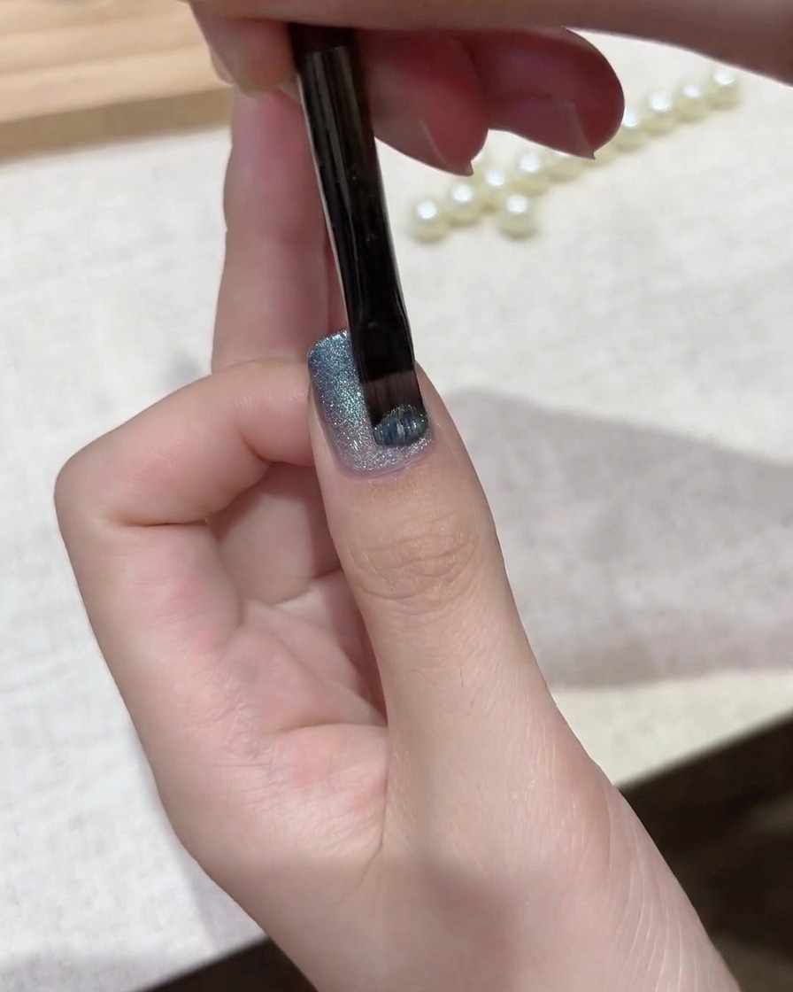 ジェルブラシがグラデーションベースの上にきらめくシルバーブルーのマグネットキャッツアイポリッシュの層を塗布している様子。