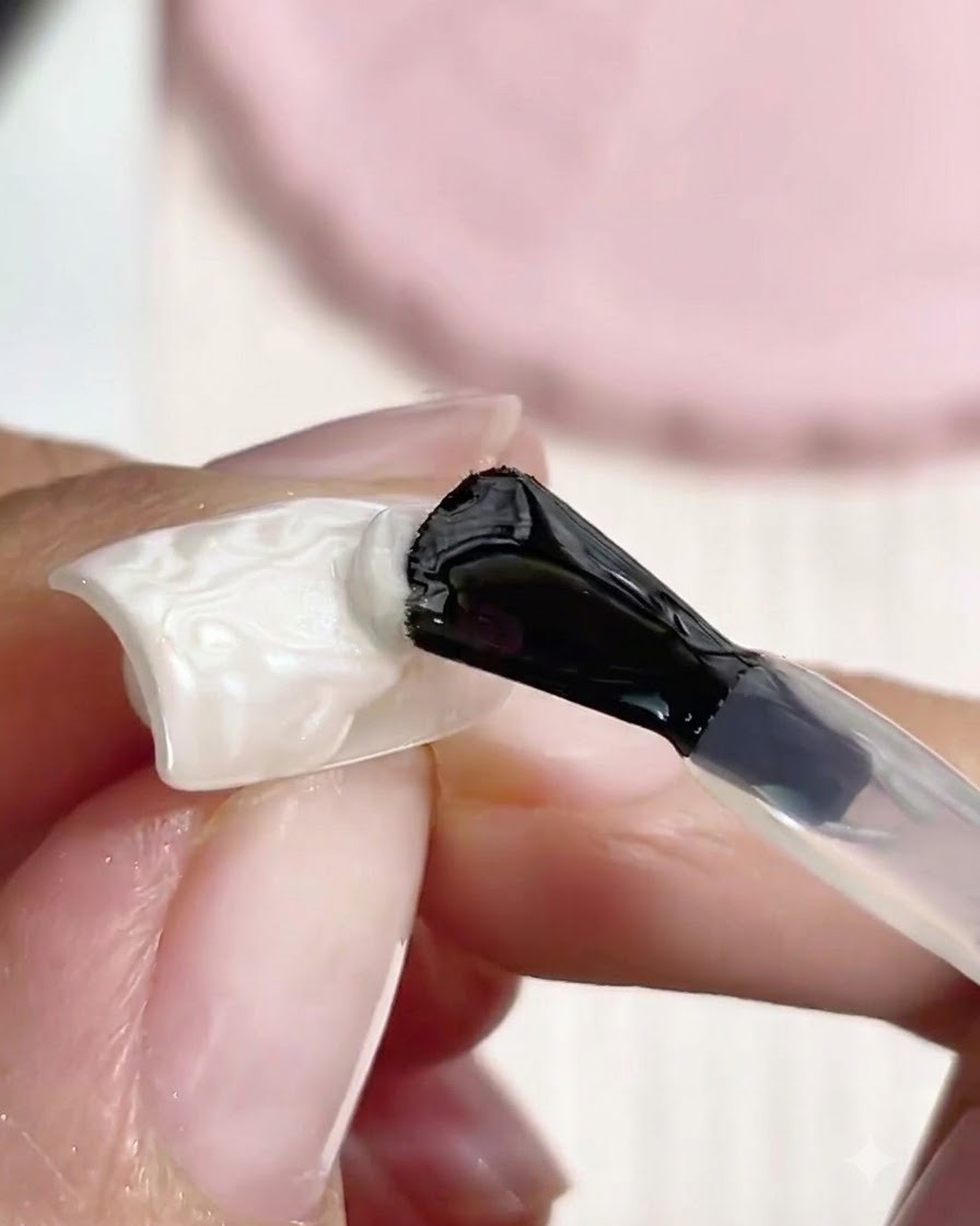 黒い柄のネイルブラシが、白いテクスチャーのある3D成形パールネイルアートの上にクリアなトップコートを塗布しているところ。