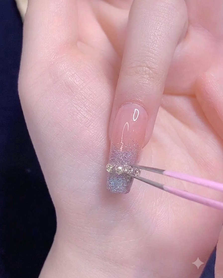 ピンクのピンセットを用いて、事前に測った小さなメタルチェーンストリップを薬指のグラデーションの先端に水平に配置している様子。
