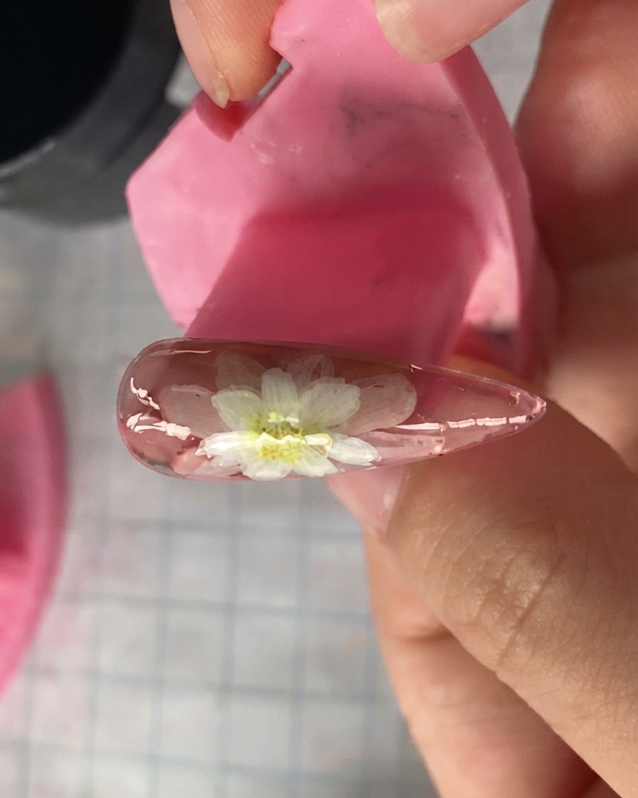 上部に描かれた重なり合う白いデイジーの花びらを示す、ピンクのネイルモデルのクローズアップ。