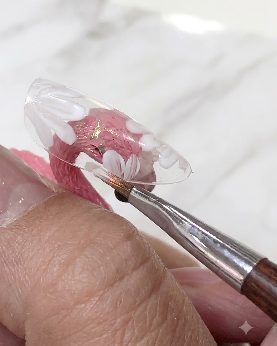 クリアネイルチップ上の描かれた白いお花の上に、トランスルーセントなピンクのジェルポリッシュのレイヤーをブラシで塗布している様子。