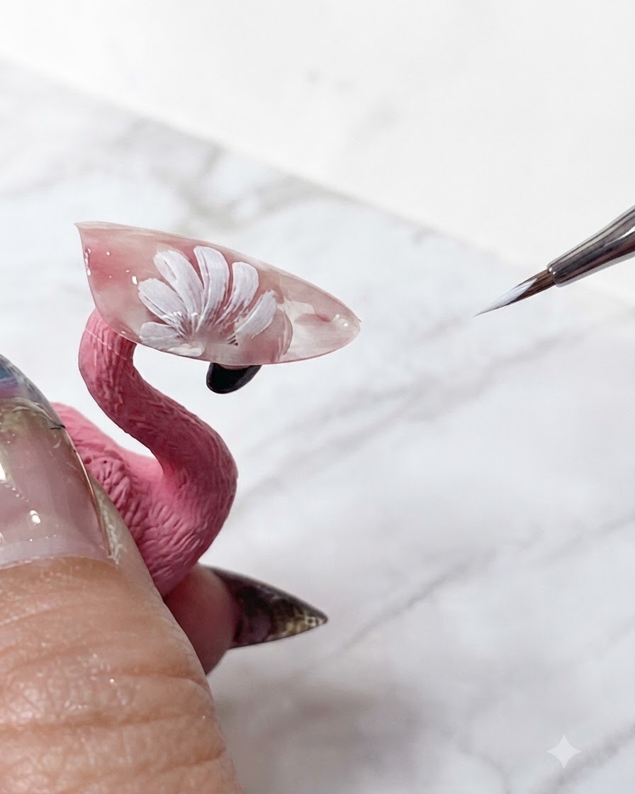 半透明のピンクのエクステンション上で、レイヤードされたデイジーの花びらのシャープな白い先端を微調整している極細ネイルアートブラシ。
