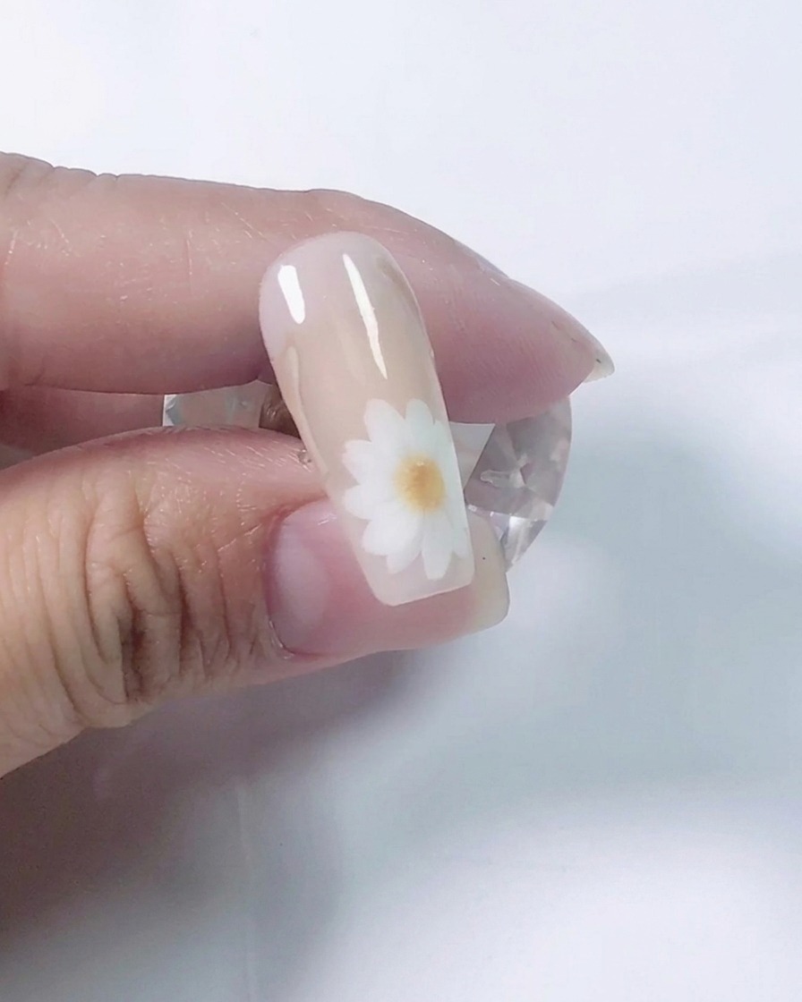 ネイリストが極細筆を使い、ヌードカラーを塗った練習用ネイルチップに白いデイジーの花びらを描いているクローズアップ写真。