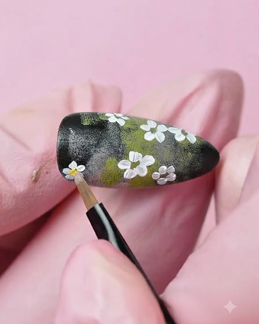 極細ディテールブラシを使用し、ネイル上に描かれた白い花の中心に黄色いジェルのドットを置いている様子。