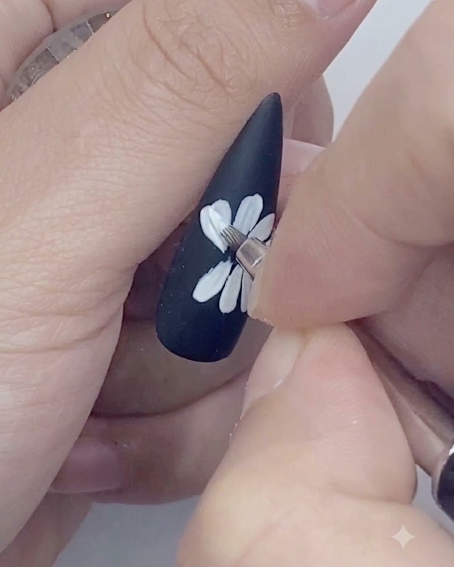 黒いネイルに描かれた白いデイジーの花びらの外側を、細いライナーブラシでトレースしている様子。