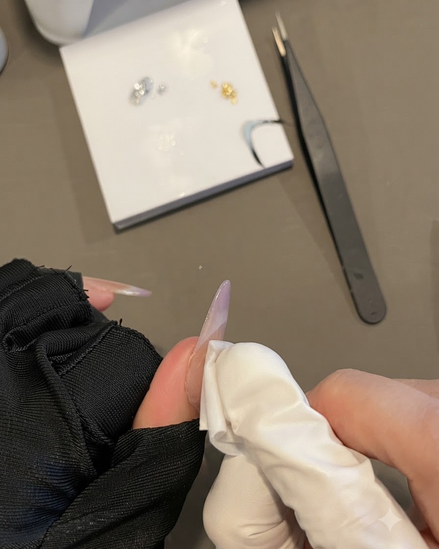 ロングオーバルネイルを持ちながら、リントフリーワイプで爪の表面を拭き取っている手袋をした手。