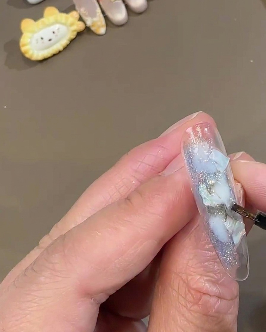 ブルーのシェルが飾られたクリアなチップの余白に、細いディテールブラシで白いスパークリングフェアリーパウダーを乗せているところ。