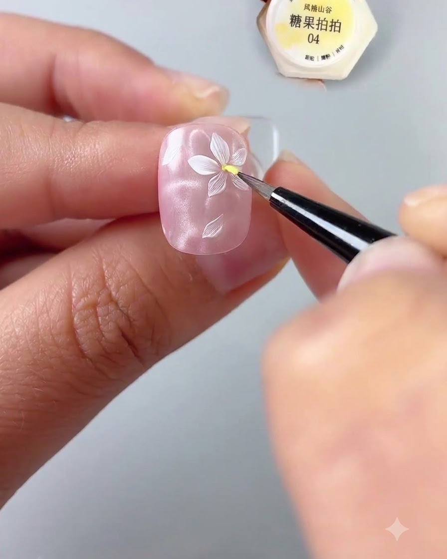 光沢のあるピンクのネイルベースに描かれた白い花の中心に、極細ディテールブラシを使って黄色のジェルのドットを置いている手元のクローズアップ。