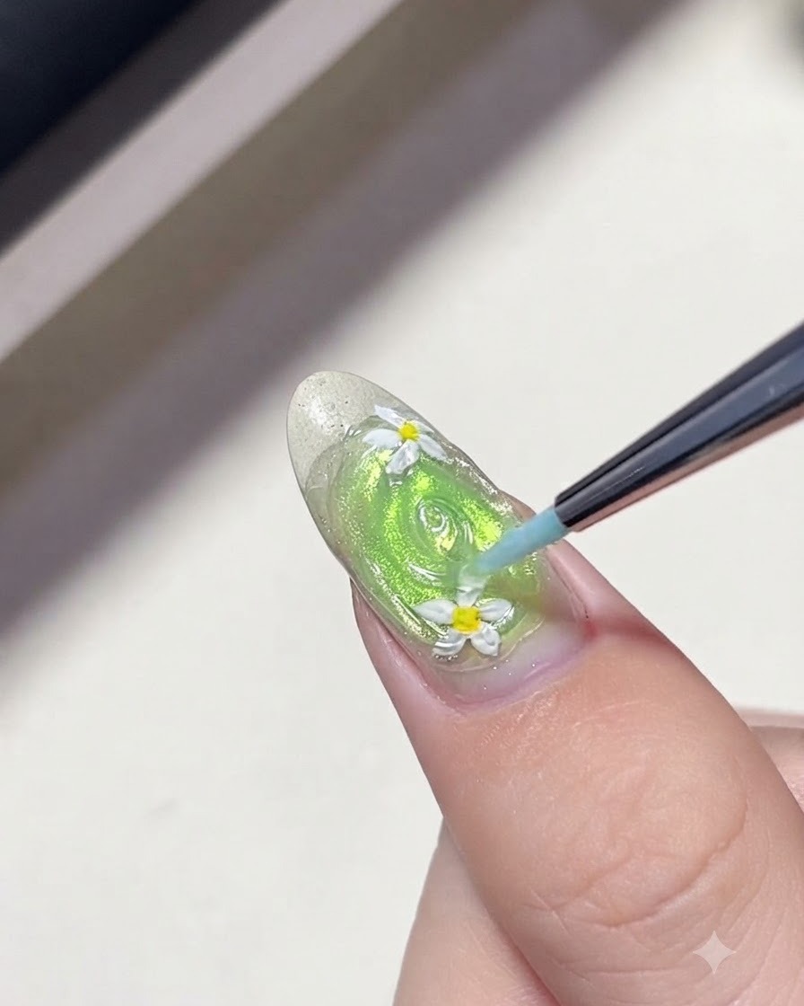 グリーンのネイルに描かれたデイジーの花のまわりに、極細のライナーブラシでクリアジェルの同心円を描いて水滴のテクスチャーを作っている様子。